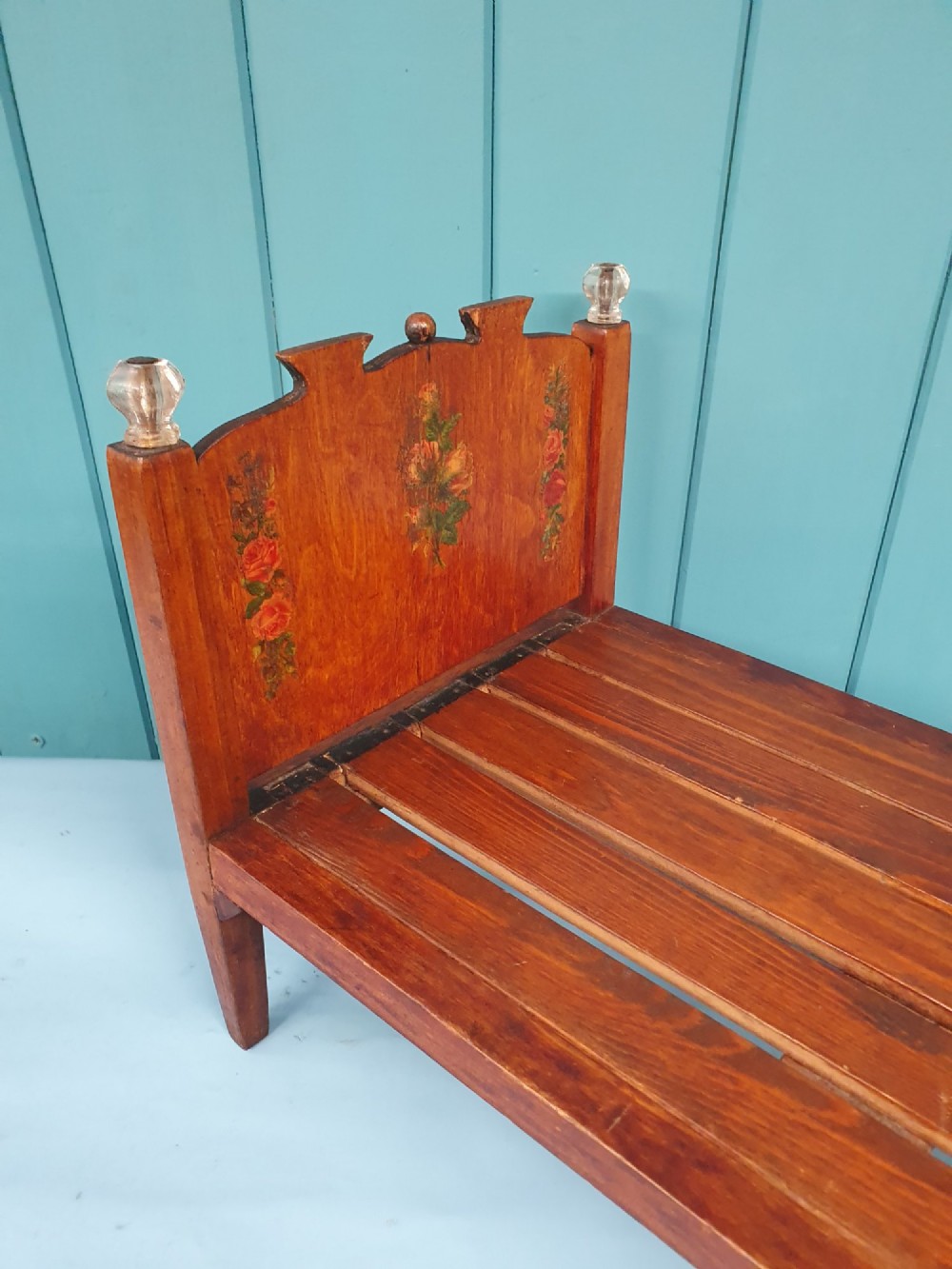 A small antique wooden doll bed with hand-painted floral motifs on the headboard and footboard, a slatted frame, and clear glass finials on four corner posts. View VII