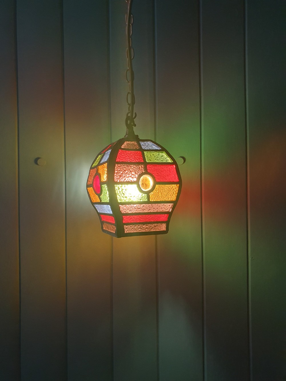 An early 20th-century leaded stained-glass porch lantern featuring geometric panels of red, green, amber, and blue glass with round glass cabochons, suspended from a brass chain. View VII