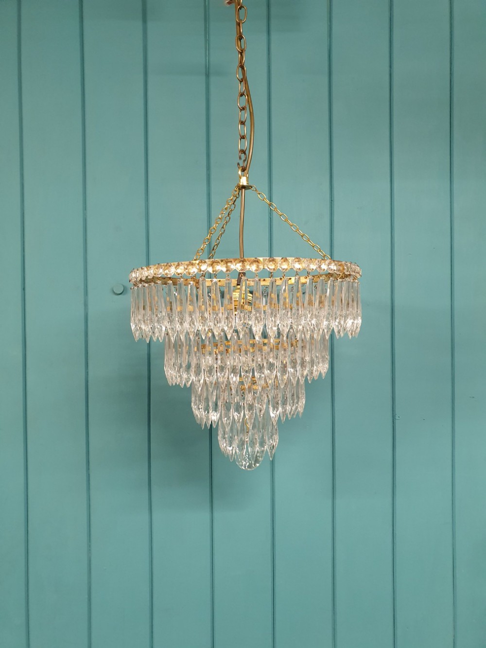 A tiered brass ceiling chandelier featuring multiple concentric rows of clear glass spike crystal prisms suspended by a chain.