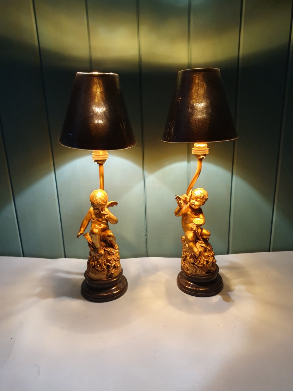 A pair of antique gilt brass table lamps featuring detailed sculpted cherub figures seated on circular dark marble bases. View II
