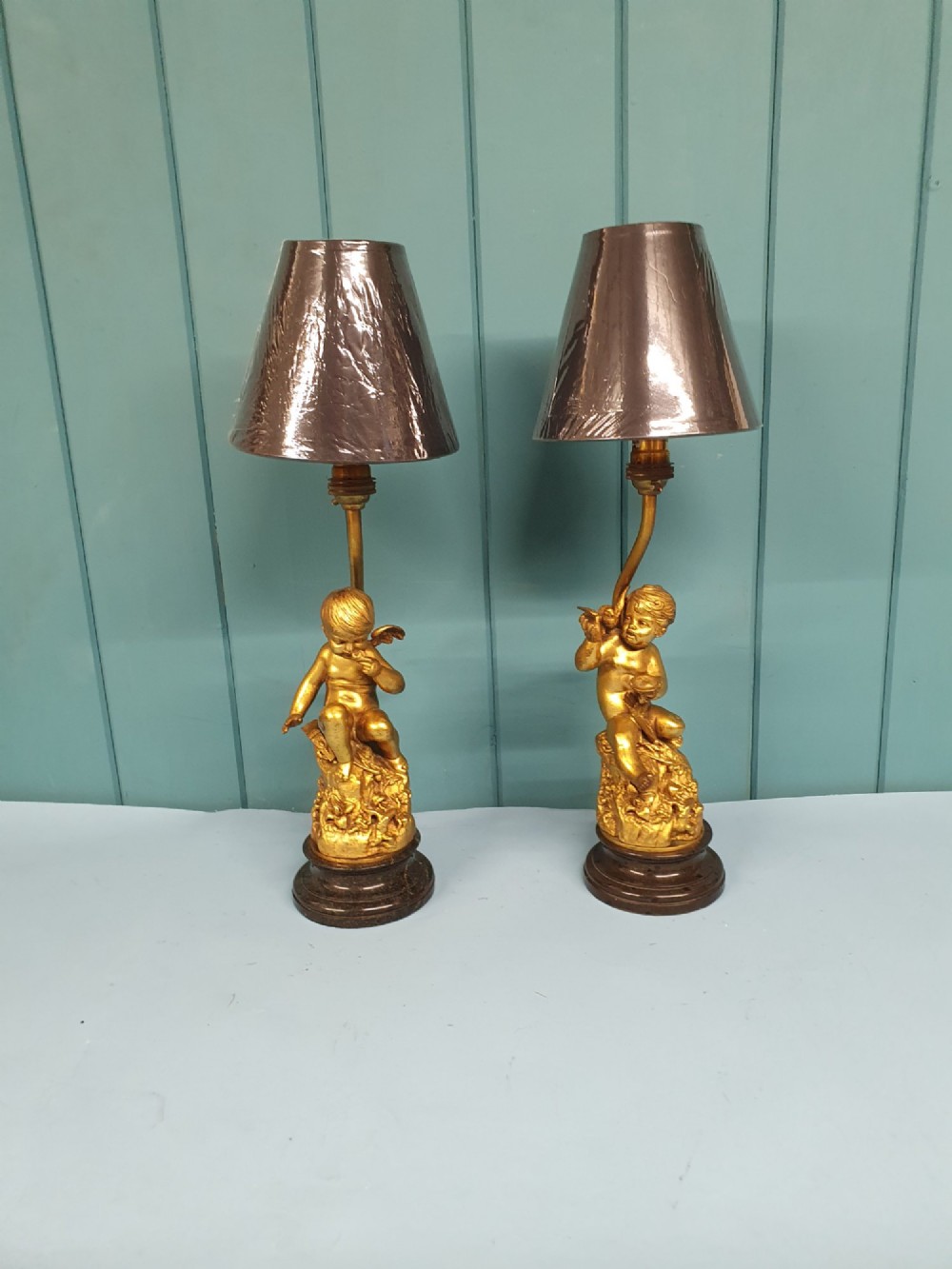 A pair of antique gilt brass table lamps featuring detailed sculpted cherub figures seated on circular dark marble bases. View VI