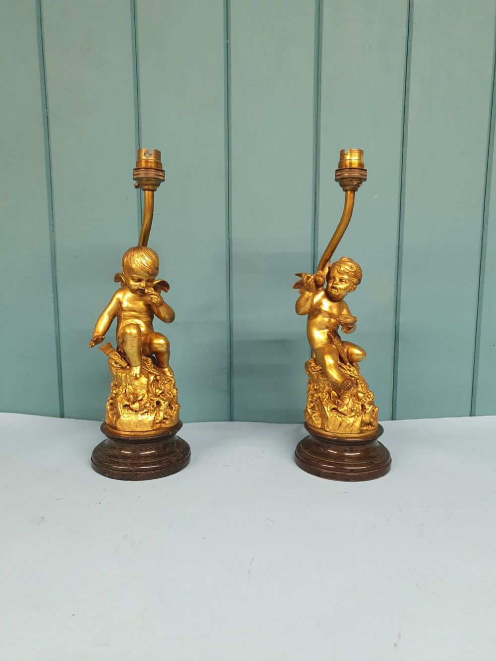 A pair of antique gilt brass table lamps featuring detailed sculpted cherub figures seated on circular dark marble bases.