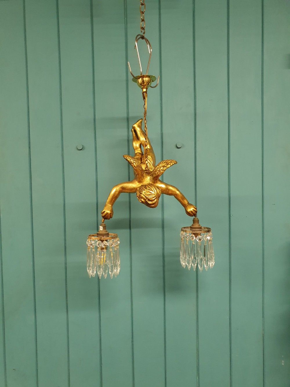 An antique gilt spelter flying winged cherub suspended from a chain, holding two light sockets decorated with cut glass crystal drops.