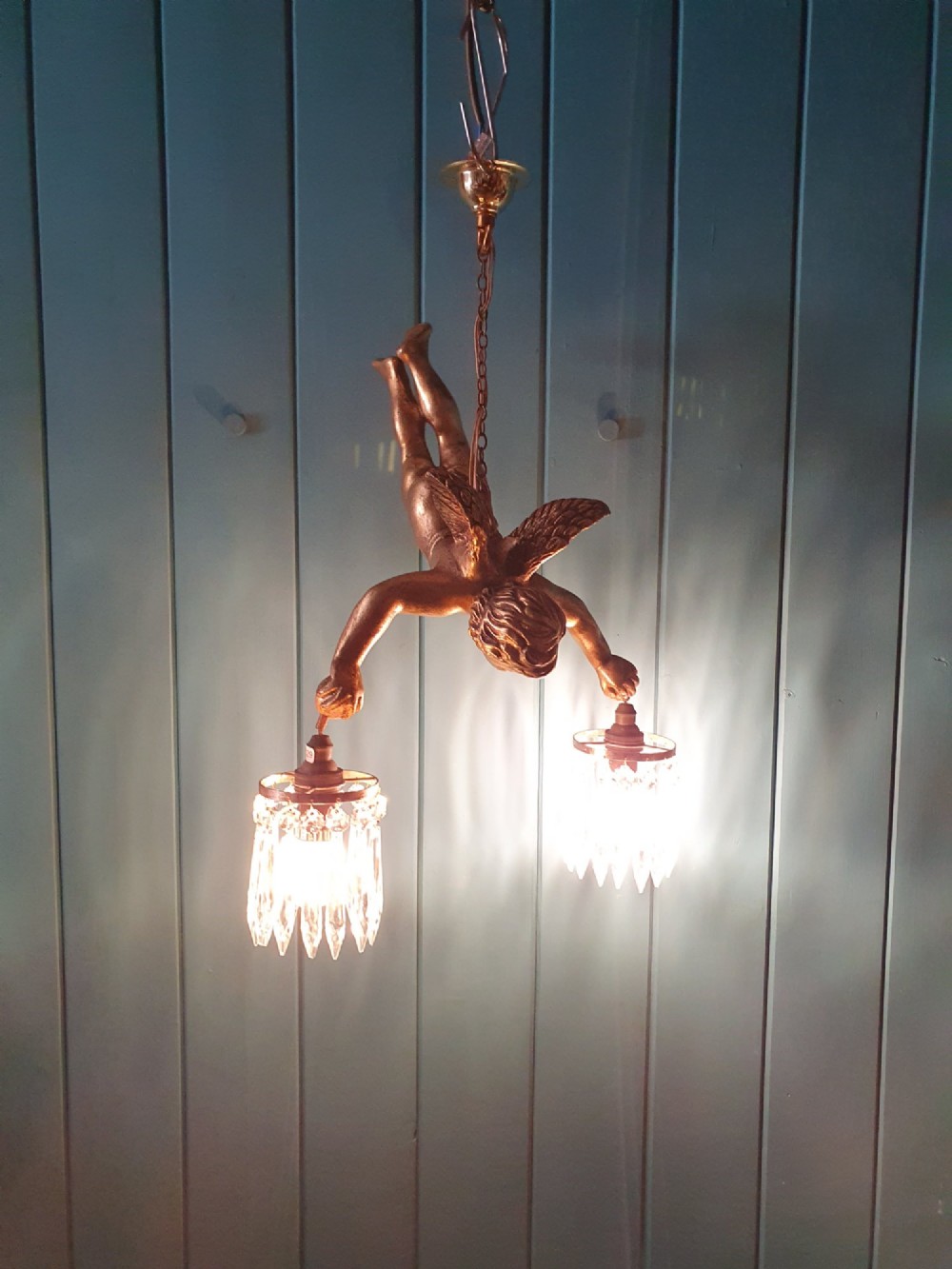 An antique gilt spelter flying winged cherub suspended from a chain, holding two light sockets decorated with cut glass crystal drops. View V