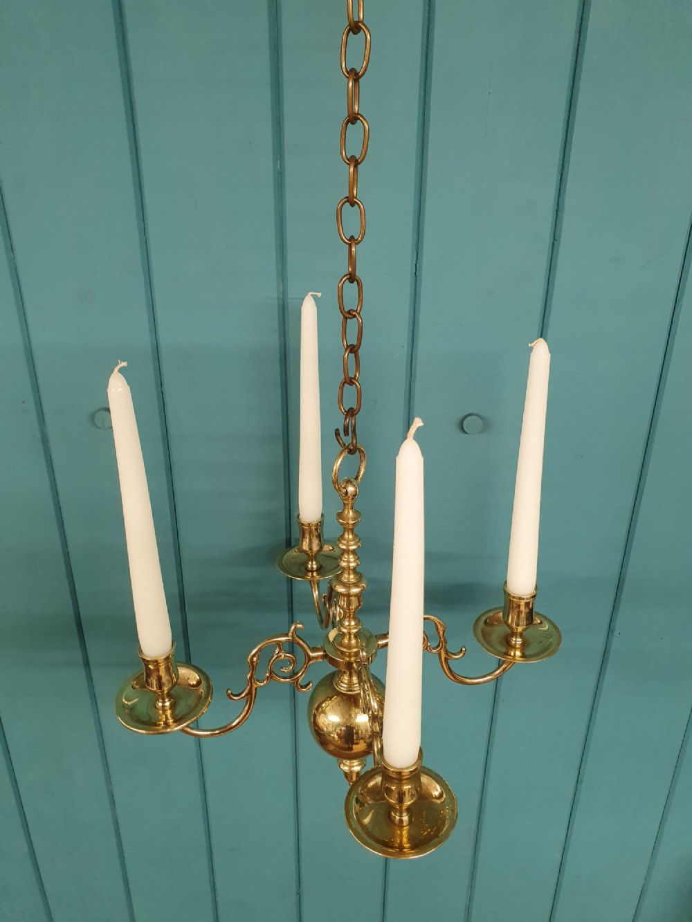 A small four-arm brass candle chandelier featuring scroll branches and a central turned column suspended from a brass chain.