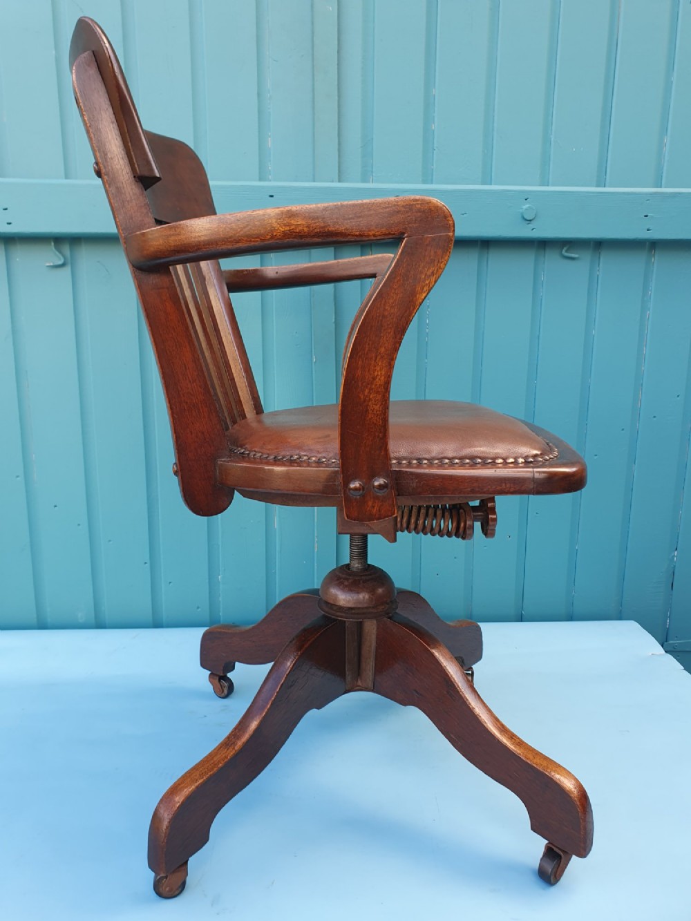 An Edwardian dark wood swivel desk chair with a slatted backrest, curved armrests, a brown leather padded seat, and four splayed legs on castors. View III