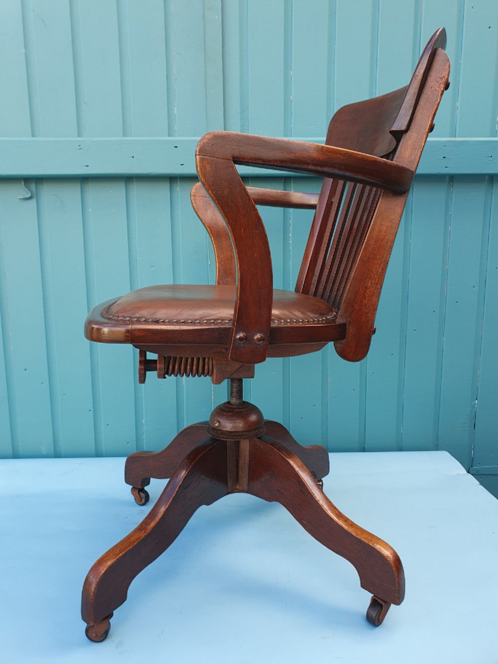 An Edwardian dark wood swivel desk chair with a slatted backrest, curved armrests, a brown leather padded seat, and four splayed legs on castors. View V