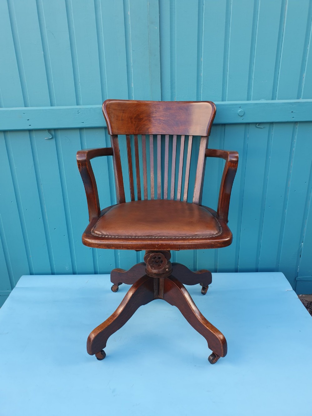 An Edwardian dark wood swivel desk chair with a slatted backrest, curved armrests, a brown leather padded seat, and four splayed legs on castors.