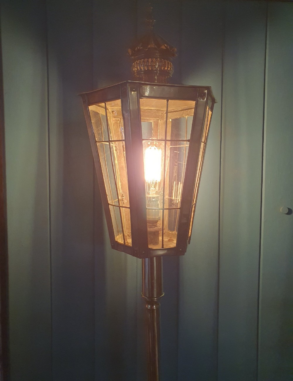 A tall, polished brass floor-standing lantern lamp featuring a hexagonal glass enclosure mounted on a slender turned stem and a circular stepped base with paw feet. View VI