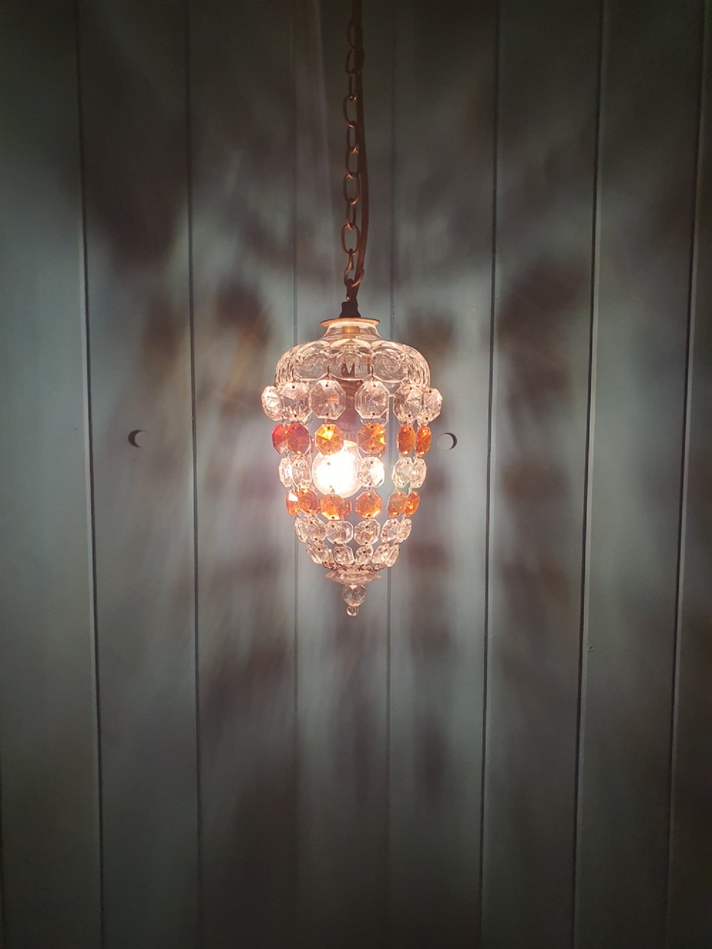An early 20th-century bag chandelier with clear and amber faceted crystal beads hanging from a brass chain against a green wooden panelled background. View II