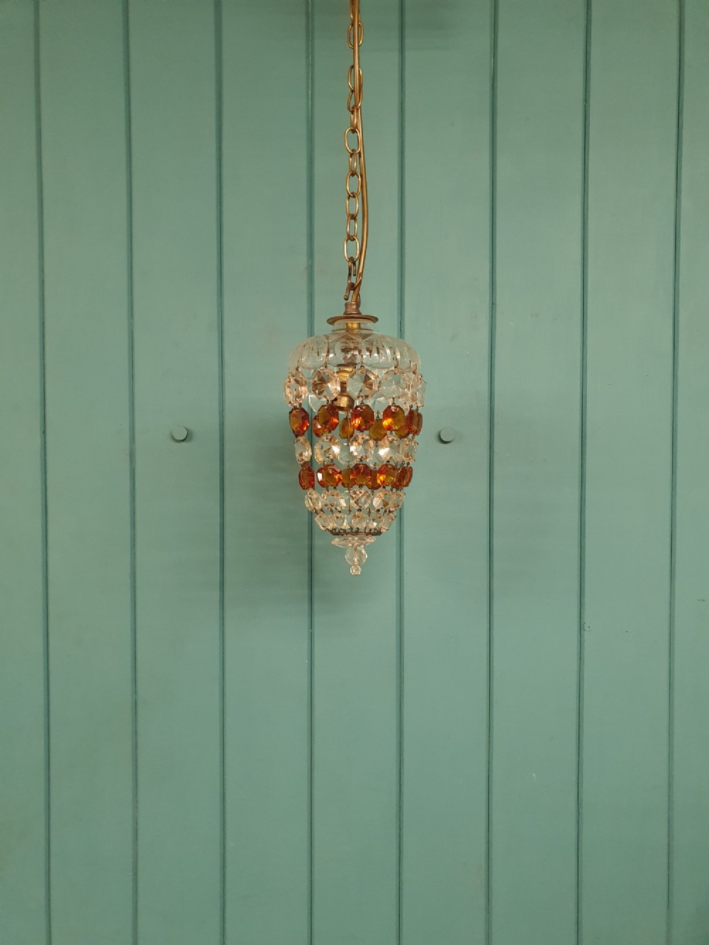 An early 20th-century bag chandelier with clear and amber faceted crystal beads hanging from a brass chain against a green wooden panelled background.