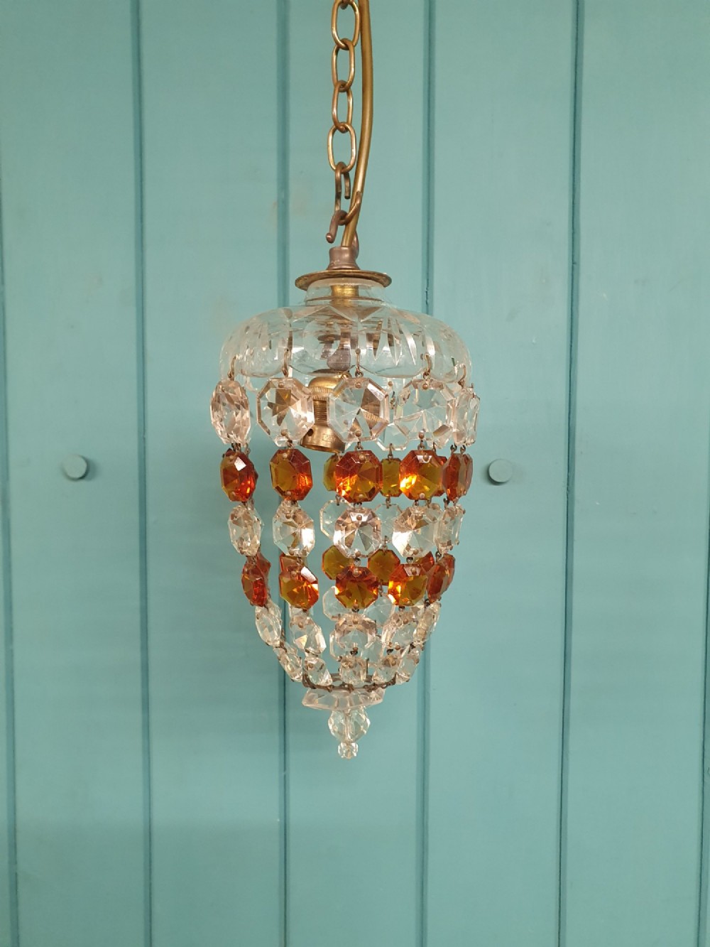 An early 20th-century bag chandelier with clear and amber faceted crystal beads hanging from a brass chain against a green wooden panelled background. View VI