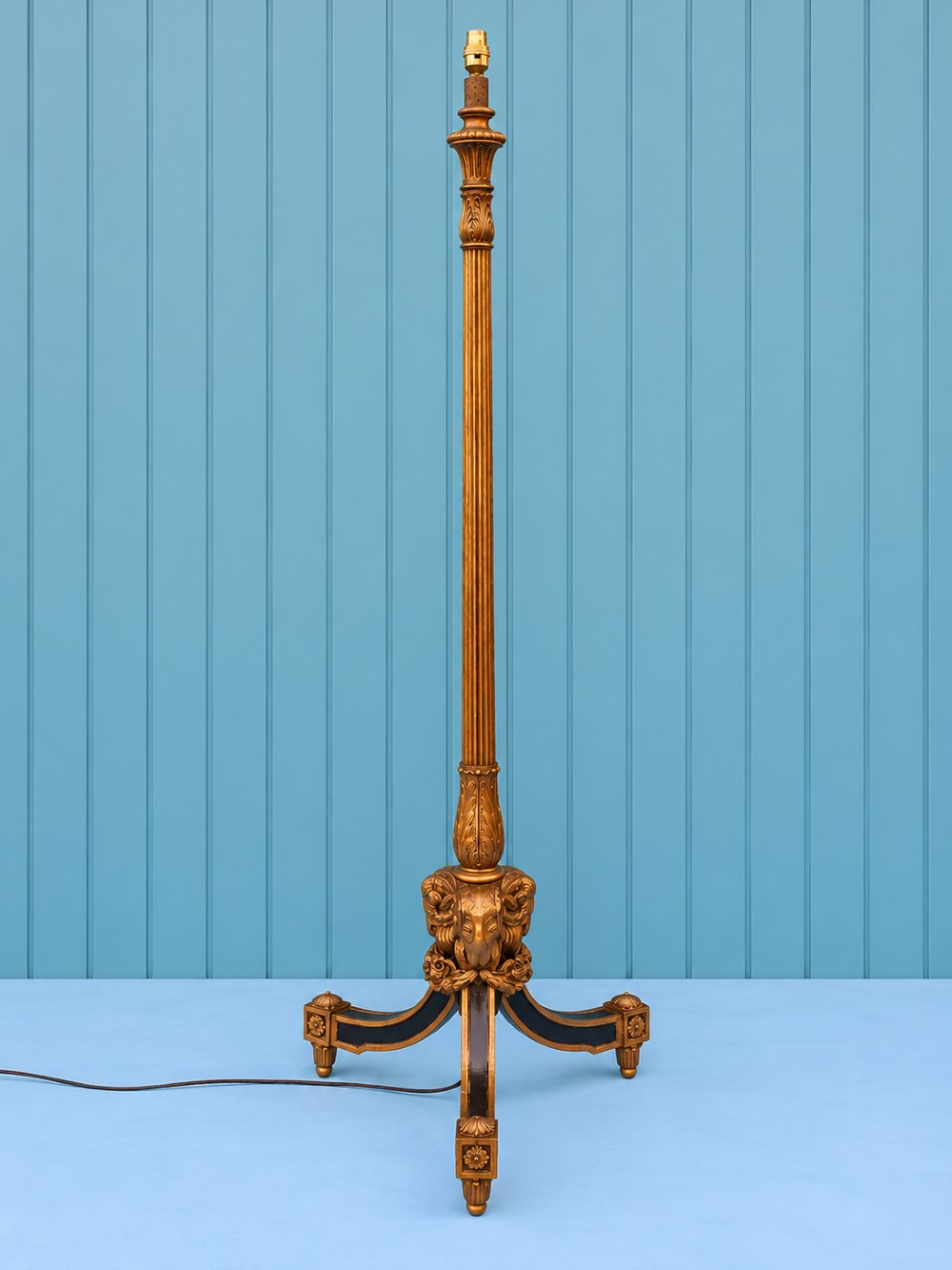 A tall antique carved giltwood standard lamp featuring a fluted column and triform base set against a blue panelled wall.