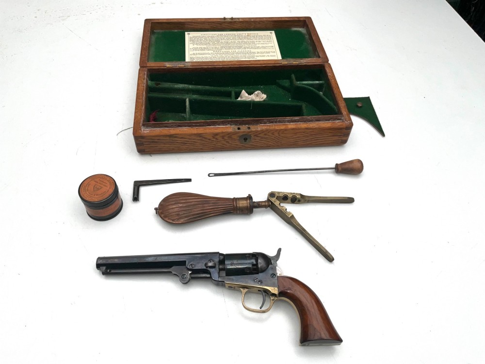 An open fitted wooden presentation case containing a .31 calibre percussion revolver, a brass powder flask, a bullet mould, and an instruction label attached to the inner lid. View V