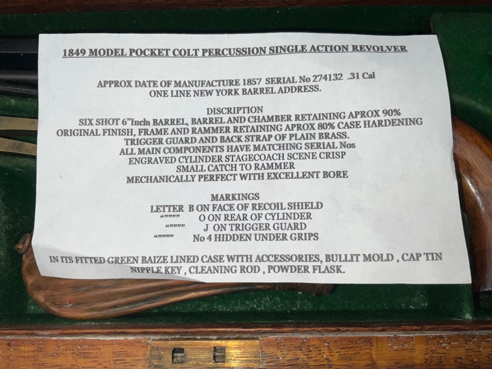 An open fitted wooden presentation case containing a .31 calibre percussion revolver, a brass powder flask, a bullet mould, and an instruction label attached to the inner lid. View XII
