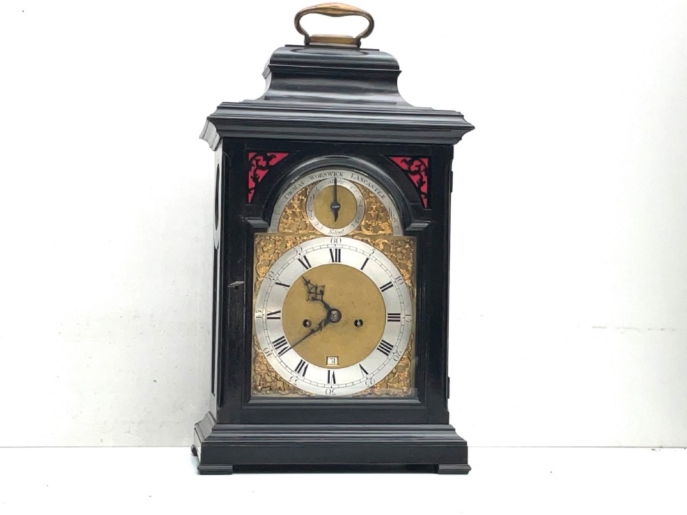 An eighteenth-century ebonised Georgian bracket clock featuring a brass carrying handle, an arched glass front door, and a brass dial with silvered chapter ring.
