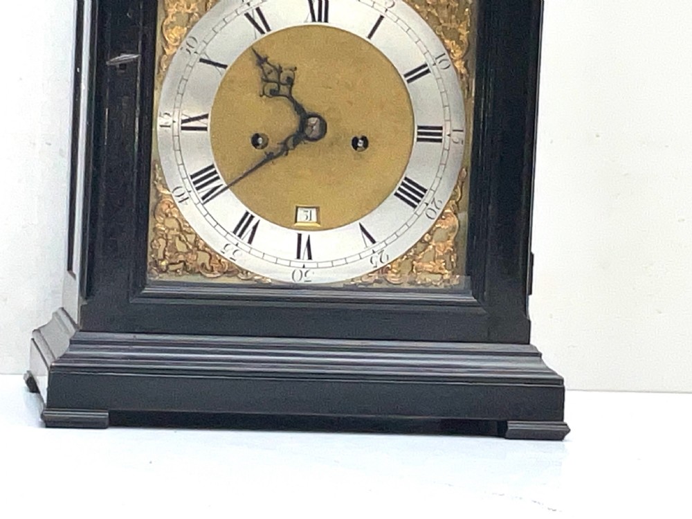 An eighteenth-century ebonised Georgian bracket clock featuring a brass carrying handle, an arched glass front door, and a brass dial with silvered chapter ring. View II