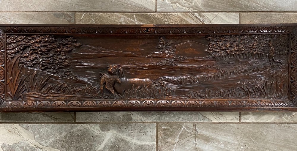 A horizontal dark walnut wood panel with a relief carving of a hunting dog in a landscape, surrounded by a carved decorative border.