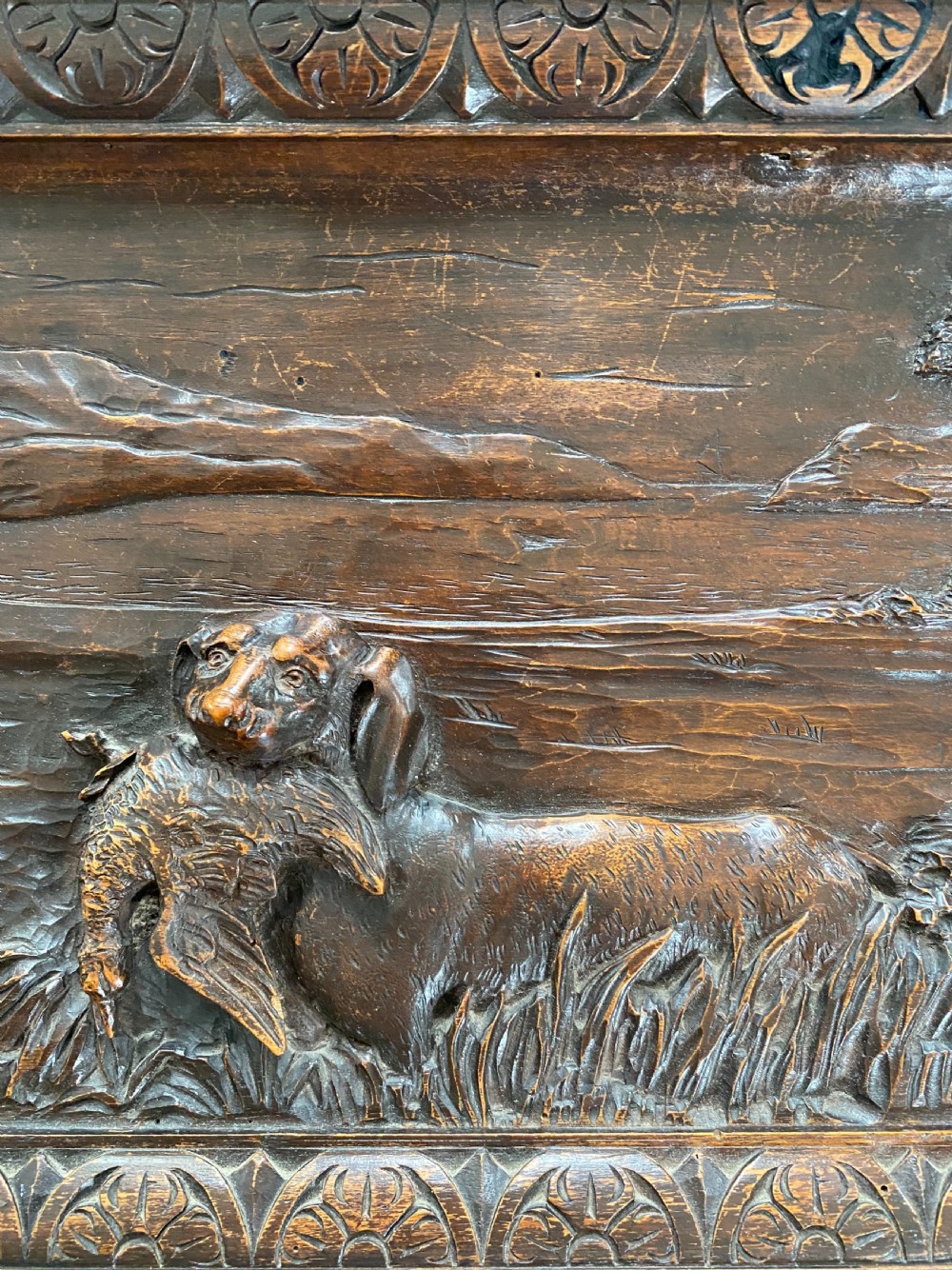 A horizontal dark walnut wood panel with a relief carving of a hunting dog in a landscape, surrounded by a carved decorative border. View III