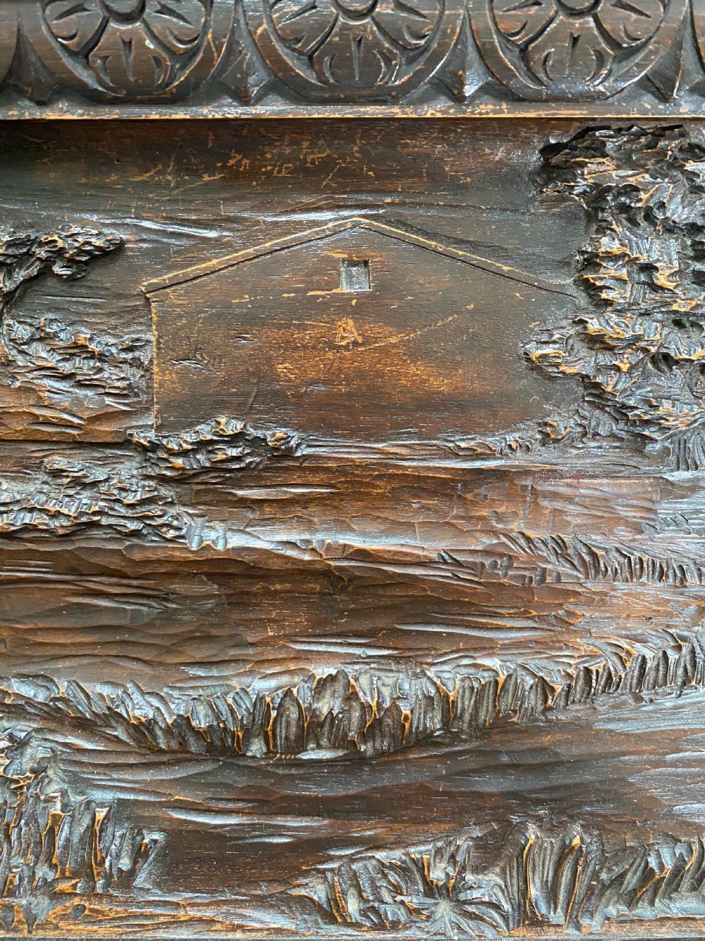 A horizontal dark walnut wood panel with a relief carving of a hunting dog in a landscape, surrounded by a carved decorative border. View IV