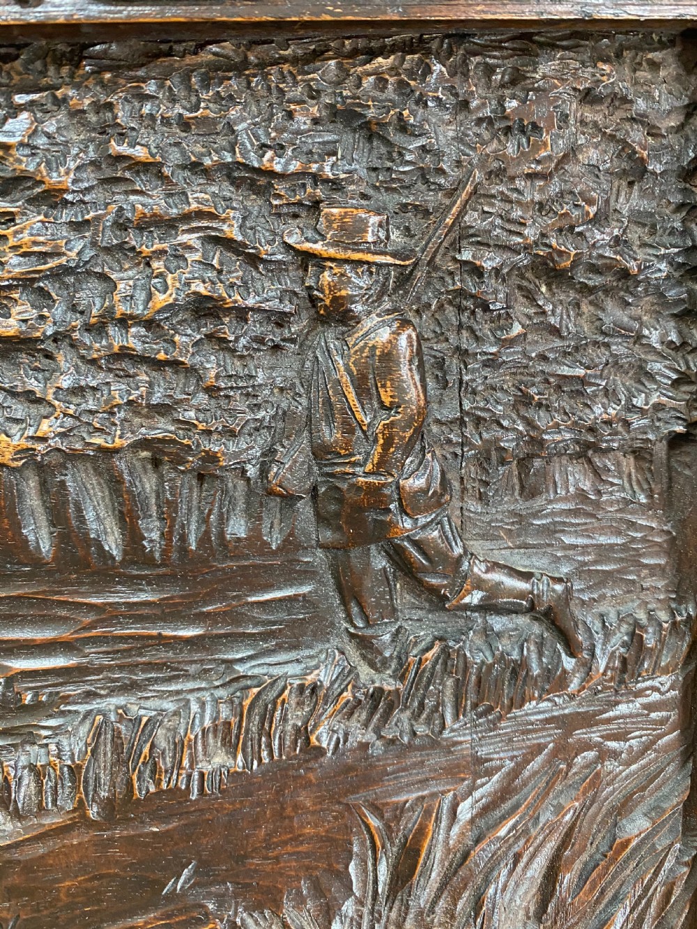 A horizontal dark walnut wood panel with a relief carving of a hunting dog in a landscape, surrounded by a carved decorative border. View V