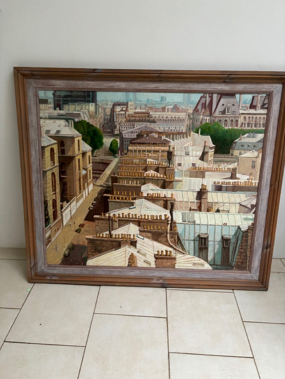 An oil painting in a dark wooden frame depicting an elevated, detailed cityscape of Parisian rooftops, chimneys, and streets.