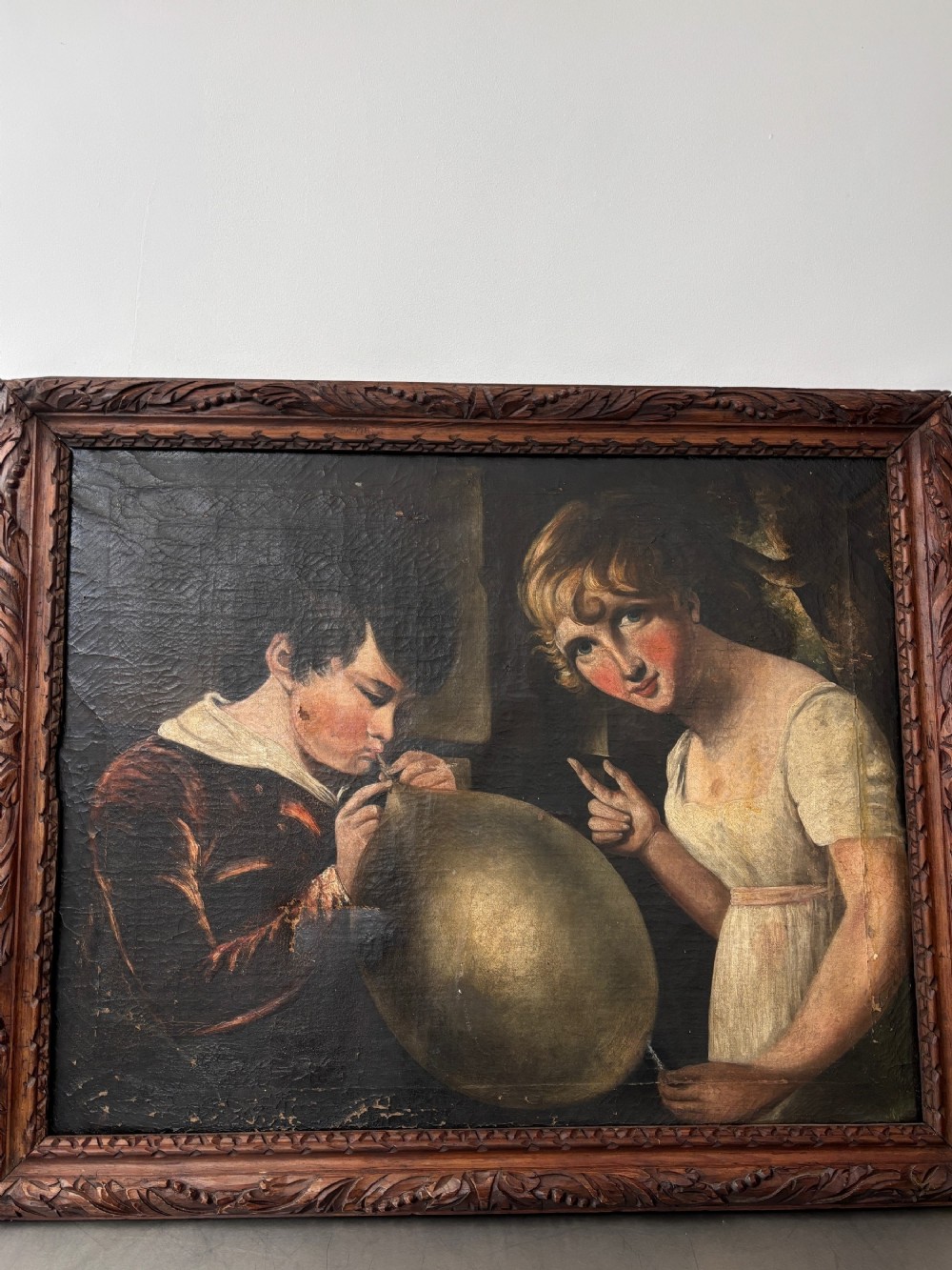 An 18th-century oil painting depicting a young boy blowing into a bladder next to a young woman, housed in a carved dark wood frame.