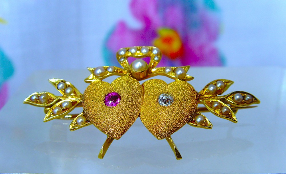 A Victorian 18ct yellow gold double heart brooch set with a ruby, a diamond, and seed pearl accents.