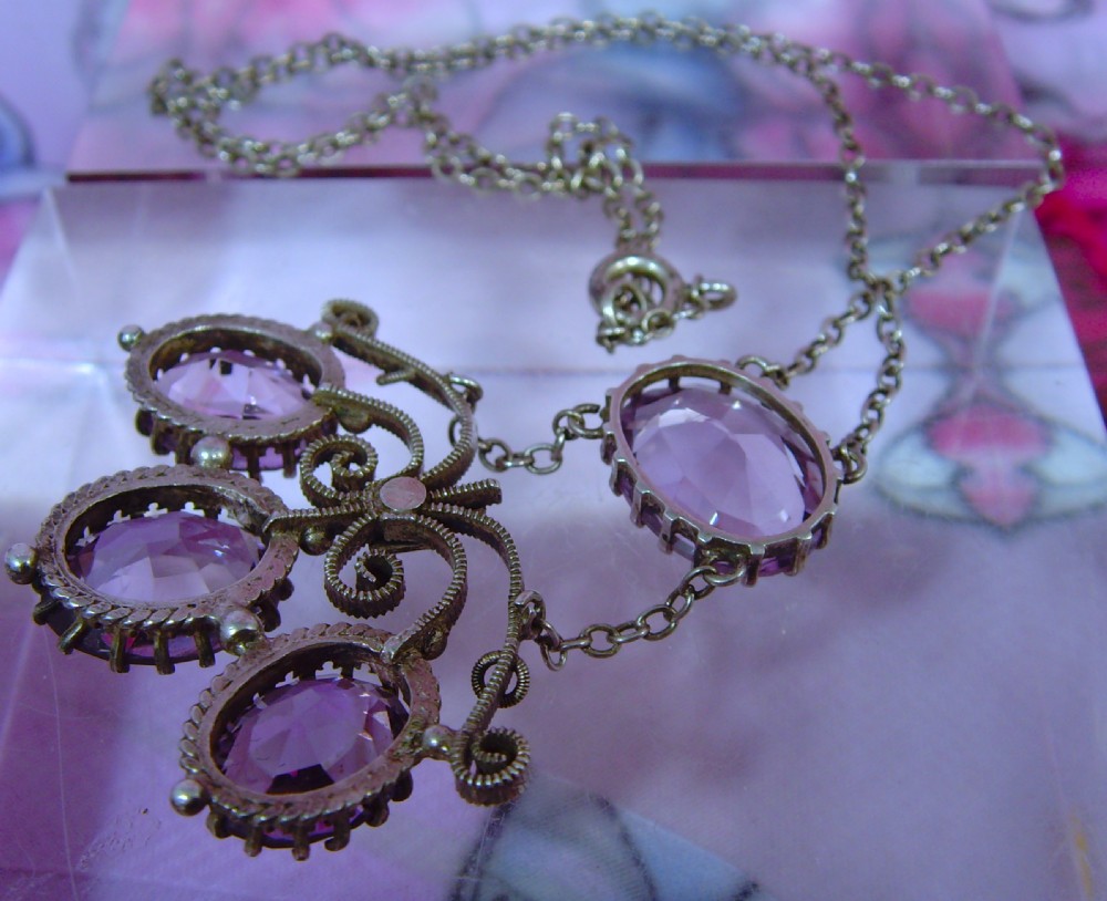 An Arts and Crafts era silver necklace featuring four faceted oval purple amethyst gemstones set within a scrollwork pendant suspended from a fine link chain. View VI