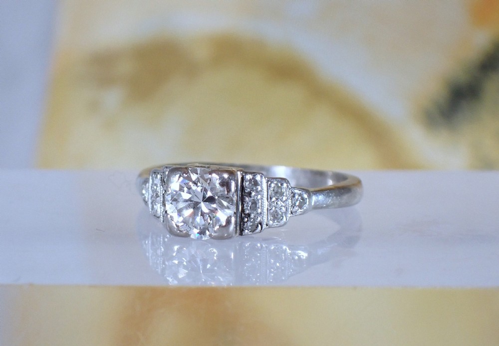 An Art Deco platinum and gold ring featuring a central round diamond set in a square frame with tiered, milgrain-detailed shoulders. View II