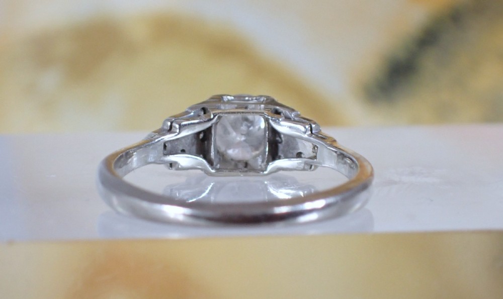 An Art Deco platinum and gold ring featuring a central round diamond set in a square frame with tiered, milgrain-detailed shoulders. View V