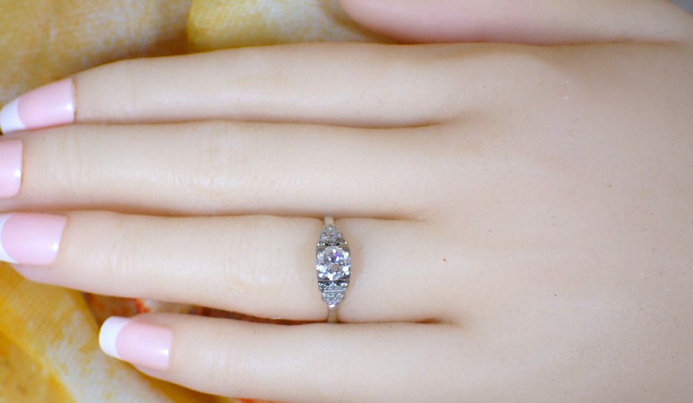 An Art Deco platinum and gold ring featuring a central round diamond set in a square frame with tiered, milgrain-detailed shoulders. View VI