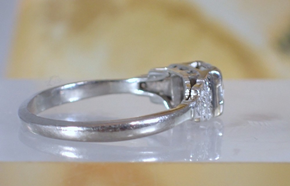 An Art Deco platinum and gold ring featuring a central round diamond set in a square frame with tiered, milgrain-detailed shoulders. View VII