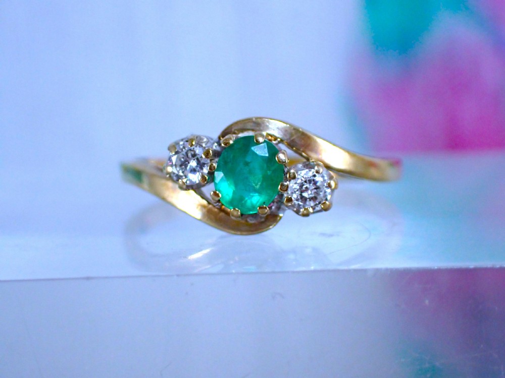 A vintage 9ct yellow gold crossover ring featuring an oval green emerald centered between two round diamonds in claw settings.
