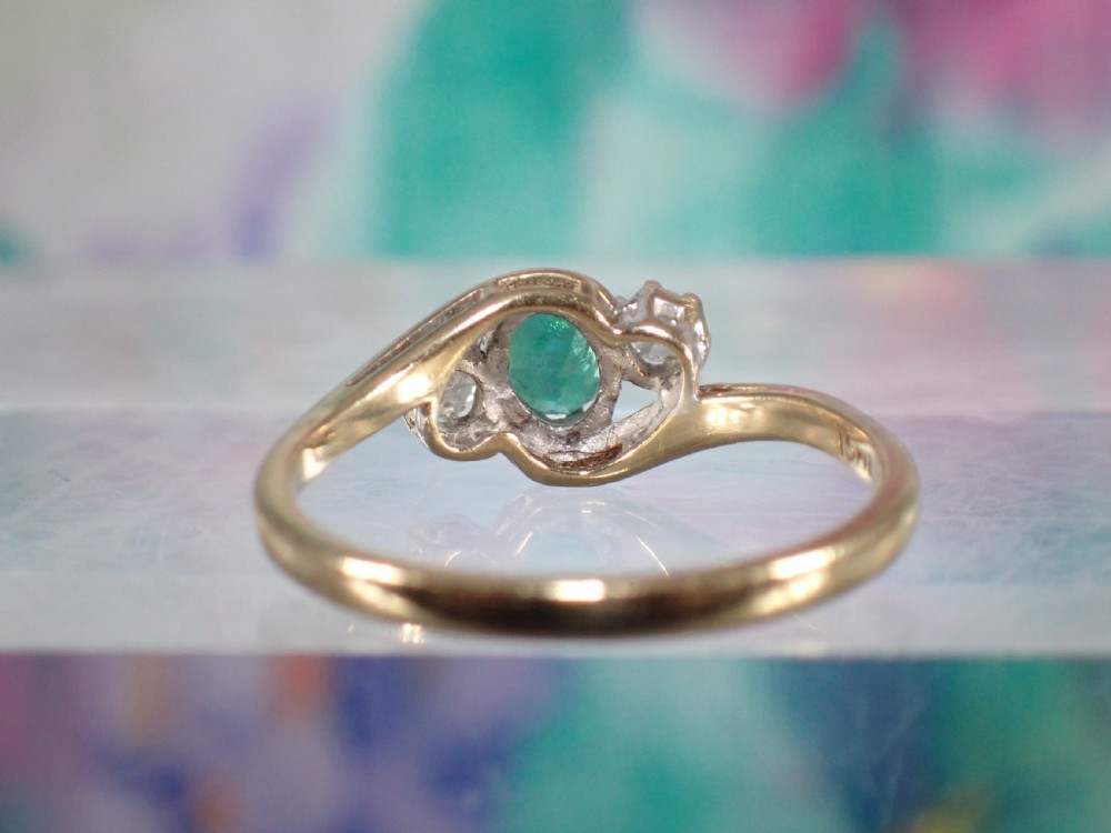 A vintage 9ct yellow gold crossover ring featuring an oval green emerald centered between two round diamonds in claw settings. View IV