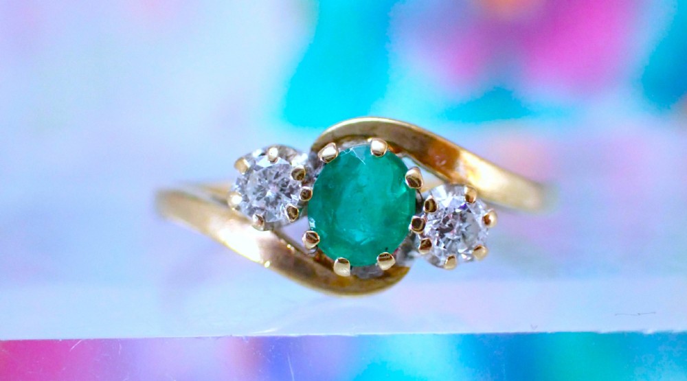 A vintage 9ct yellow gold crossover ring featuring an oval green emerald centered between two round diamonds in claw settings. View V