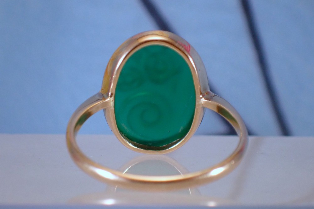 An Art Deco gold ring set with an oval green chalcedony gemstone featuring low-relief floral and scroll carving. View IX