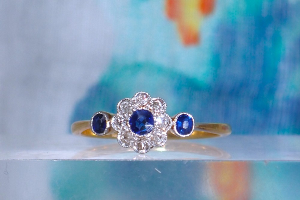 An Art Deco eighteen-carat gold ring featuring a central blue sapphire surrounded by diamonds in a daisy cluster setting, flanked by two blue sapphires on a thin yellow gold band.