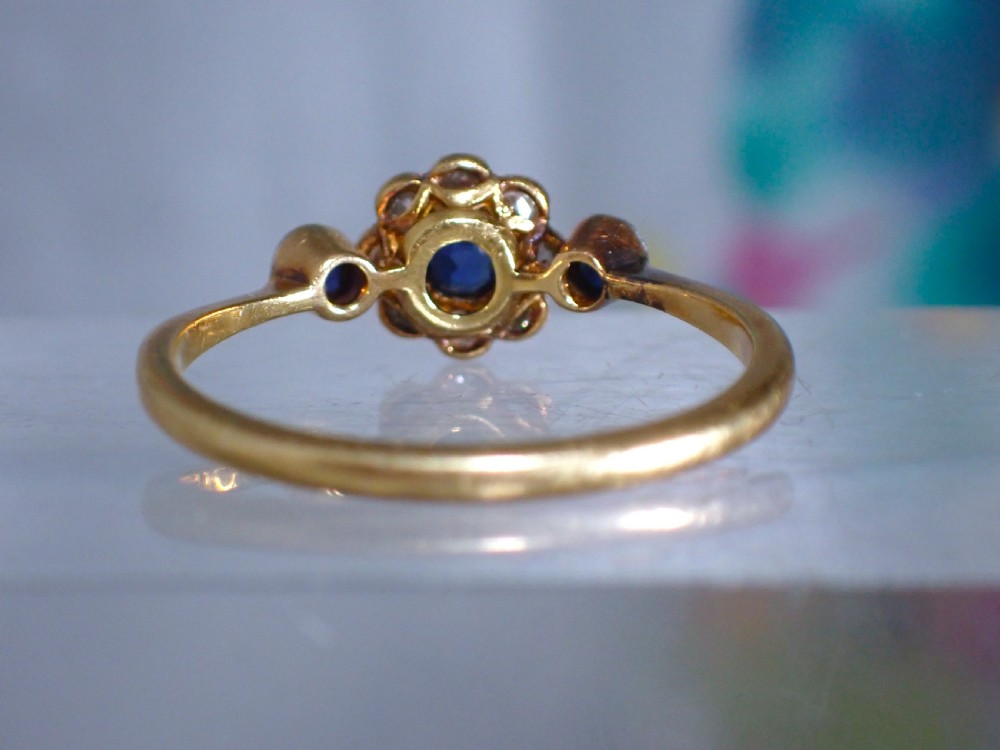 An Art Deco eighteen-carat gold ring featuring a central blue sapphire surrounded by diamonds in a daisy cluster setting, flanked by two blue sapphires on a thin yellow gold band. View IV