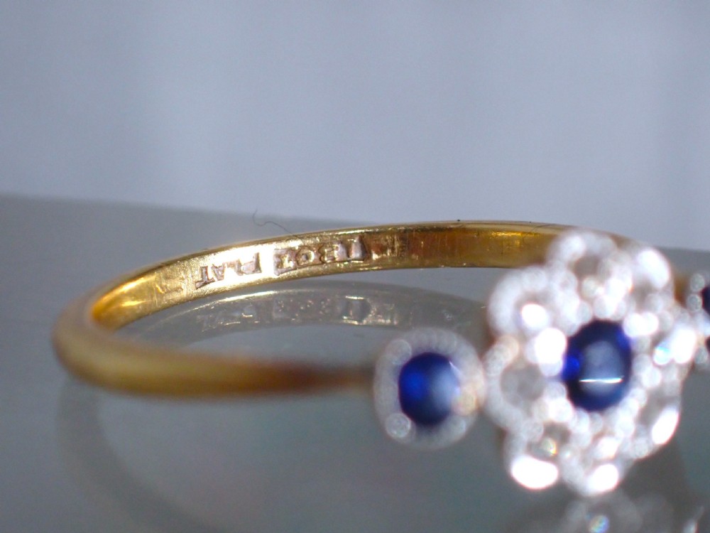 An Art Deco eighteen-carat gold ring featuring a central blue sapphire surrounded by diamonds in a daisy cluster setting, flanked by two blue sapphires on a thin yellow gold band. View VII
