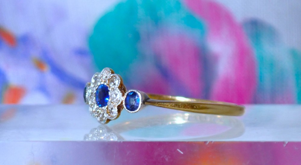 An Art Deco eighteen-carat gold ring featuring a central blue sapphire surrounded by diamonds in a daisy cluster setting, flanked by two blue sapphires on a thin yellow gold band. View VIII