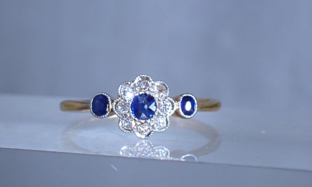 An Art Deco eighteen-carat gold ring featuring a central blue sapphire surrounded by diamonds in a daisy cluster setting, flanked by two blue sapphires on a thin yellow gold band. View X