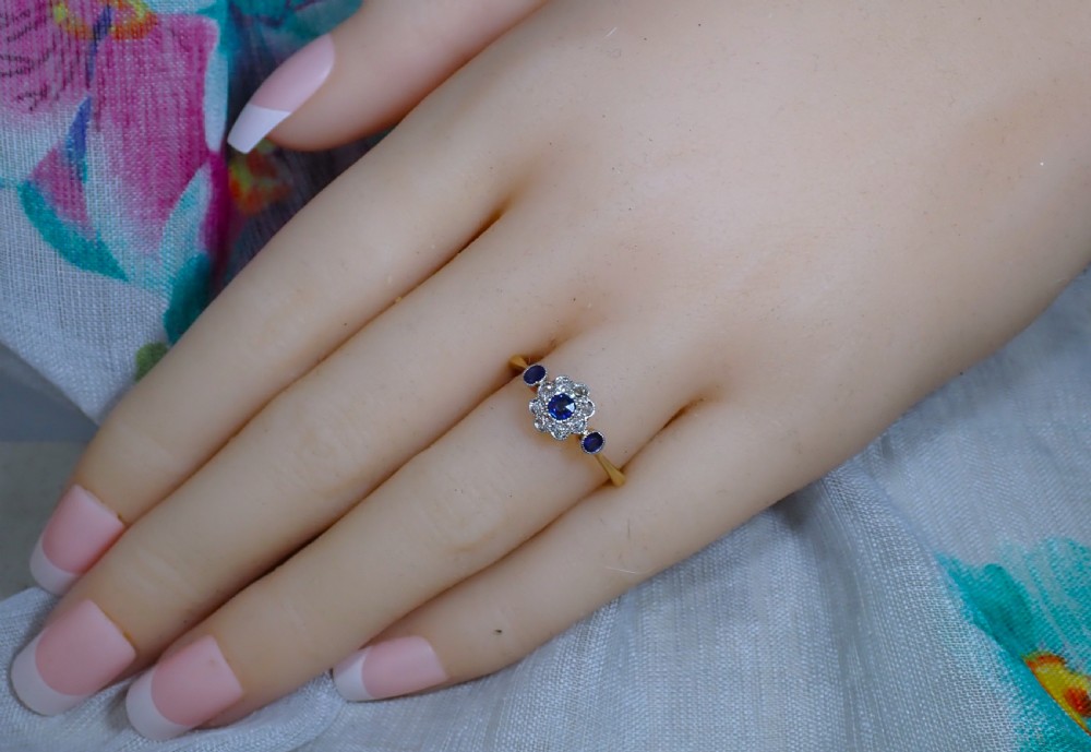 An Art Deco eighteen-carat gold ring featuring a central blue sapphire surrounded by diamonds in a daisy cluster setting, flanked by two blue sapphires on a thin yellow gold band. View XI