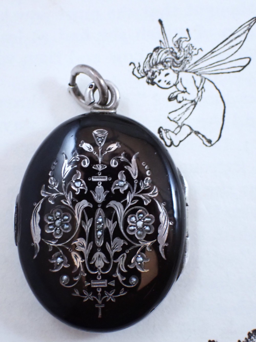 An oval Victorian black enamel locket with silver foliate inlay set with seed pearls, featuring a silver suspension bail.