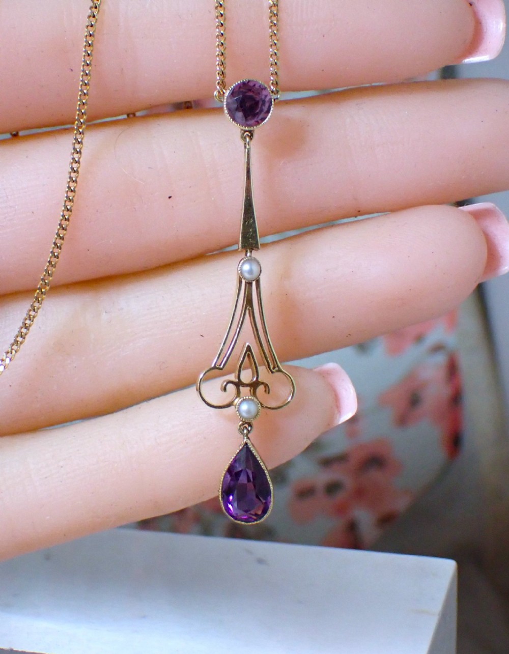 An Art Nouveau 15ct gold pendant featuring openwork scrolling metalwork set with a round amethyst at the top, two seed pearls, and a pear-cut amethyst drop suspended from a fine chain.