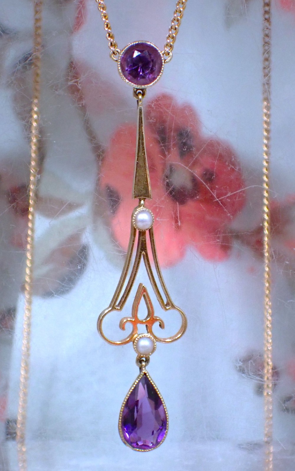 An Art Nouveau 15ct gold pendant featuring openwork scrolling metalwork set with a round amethyst at the top, two seed pearls, and a pear-cut amethyst drop suspended from a fine chain. View II