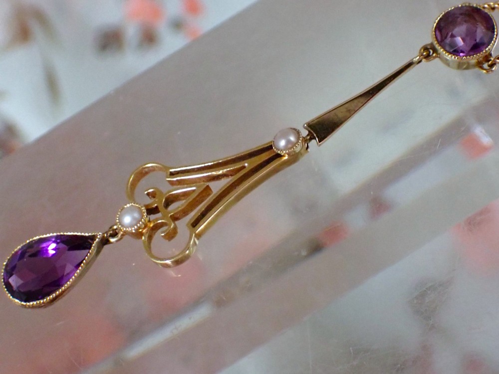 An Art Nouveau 15ct gold pendant featuring openwork scrolling metalwork set with a round amethyst at the top, two seed pearls, and a pear-cut amethyst drop suspended from a fine chain. View IV