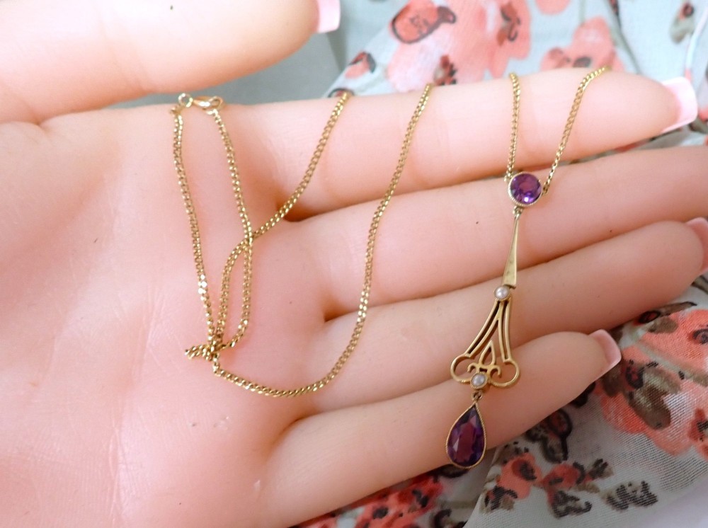 An Art Nouveau 15ct gold pendant featuring openwork scrolling metalwork set with a round amethyst at the top, two seed pearls, and a pear-cut amethyst drop suspended from a fine chain. View V