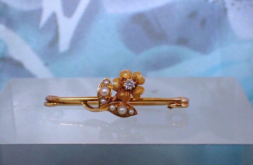 A Victorian 15ct gold bar brooch featuring a central textured flower set with a diamond and foliage set with seed pearls. View II