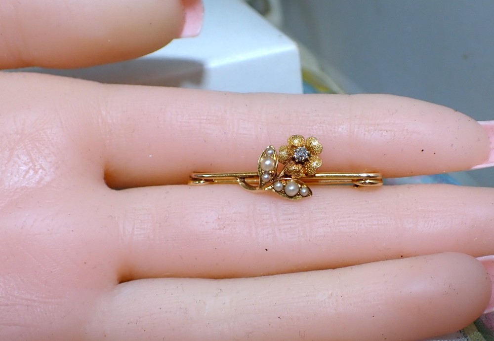 A Victorian 15ct gold bar brooch featuring a central textured flower set with a diamond and foliage set with seed pearls. View IV